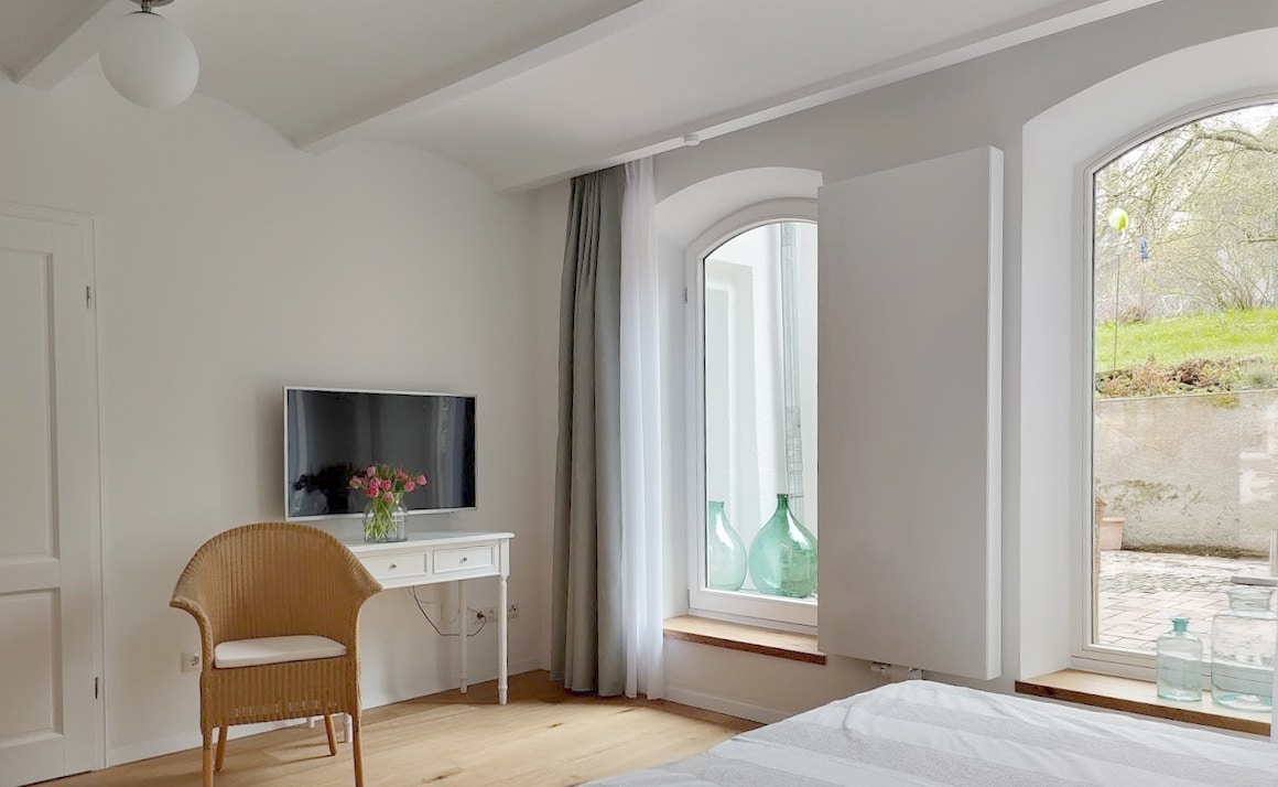 A bright room features two large windows allowing natural light to illuminate the space. A small table is positioned near the window with a vase of flowers. A comfortable chair is placed beside a TV mounted on the wall, creating a cozy seating area.