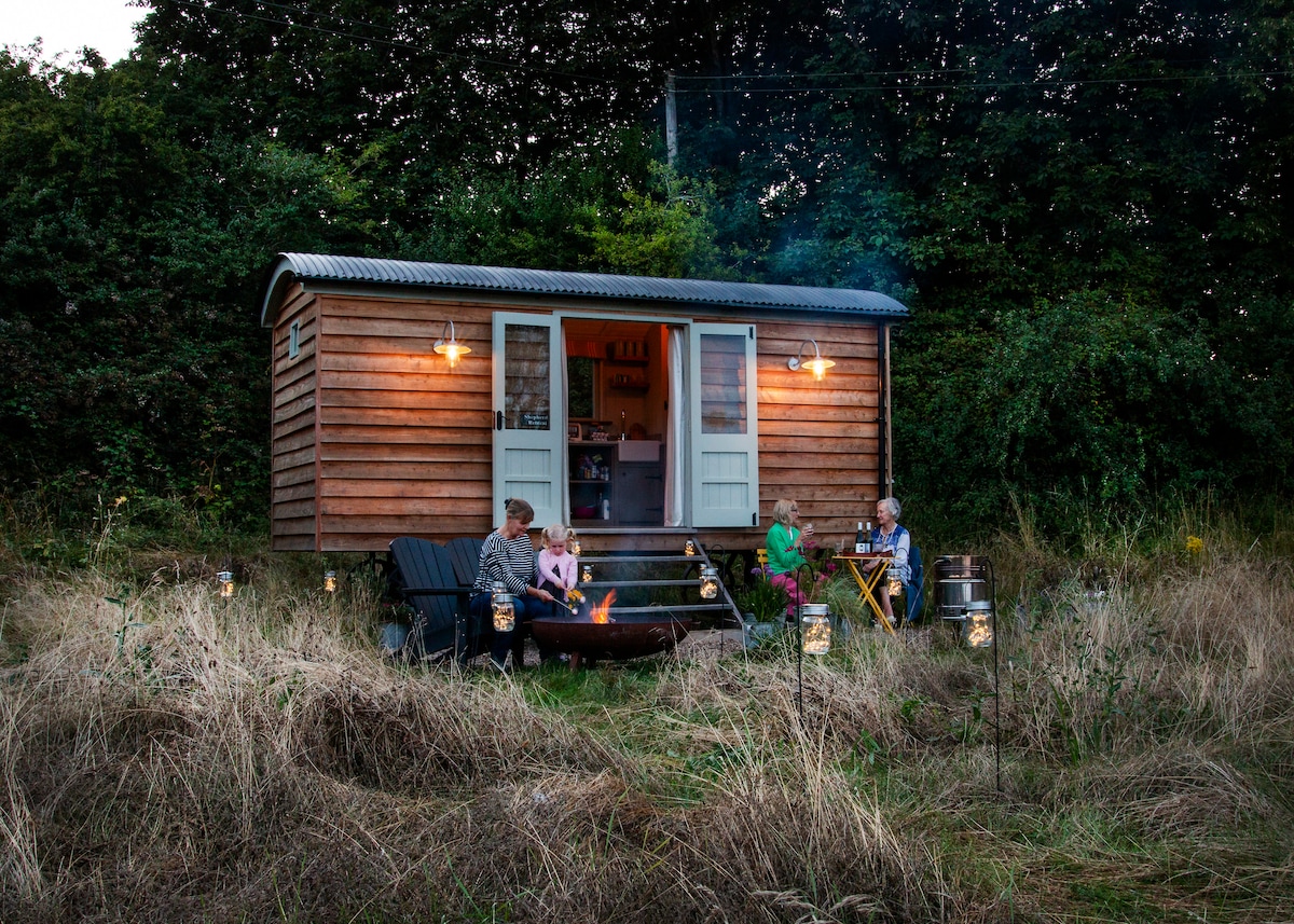 A cozy shepherd's hut is shown, featuring a wooden exterior under a twilight sky. An inviting fire pit is surrounded by seating, where four individuals enjoy a gathering. Soft lighting from the hut's exterior complements the natural surroundings.