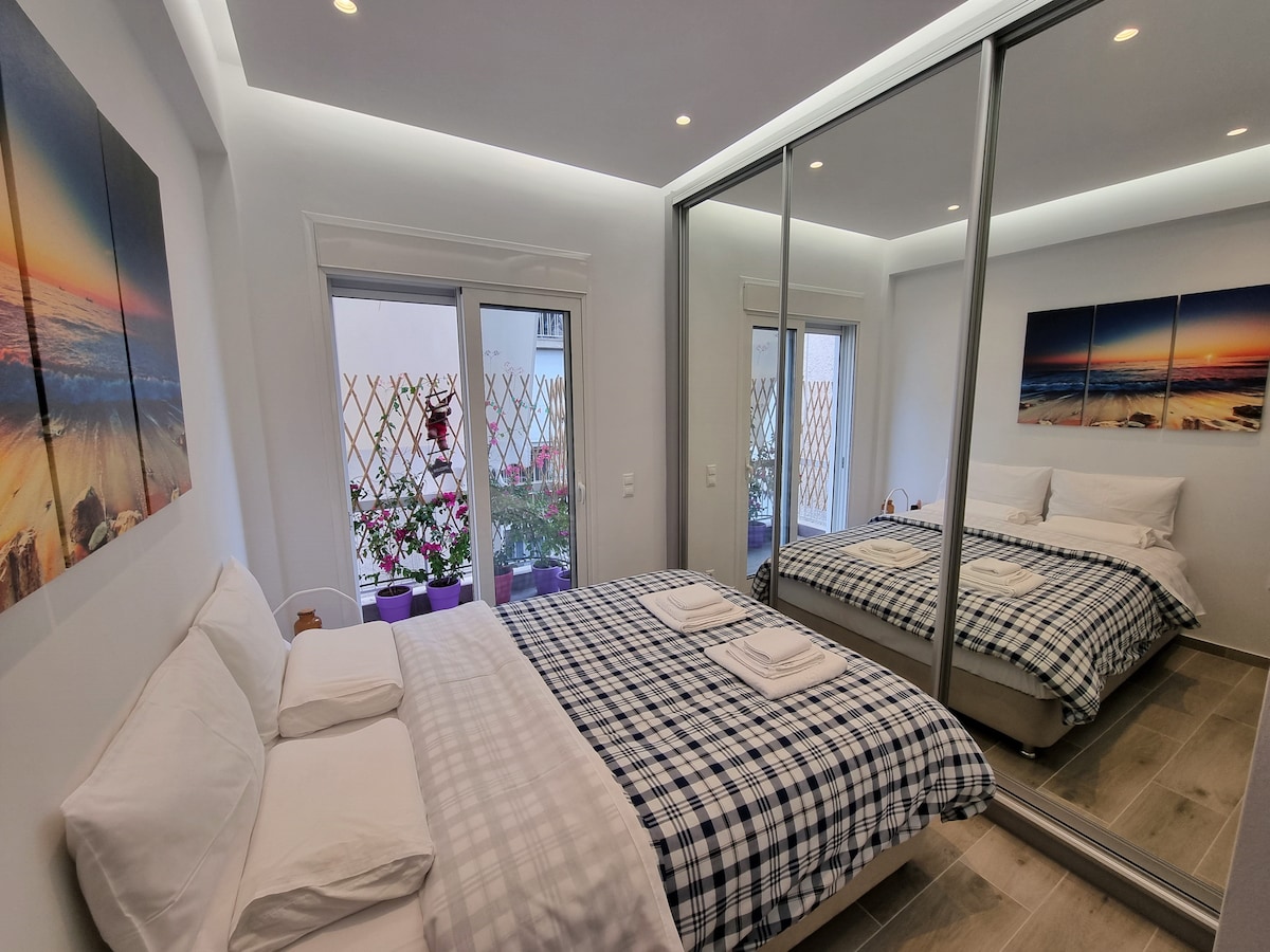 ห้องนอนพร้อมกระจกขนาดใหญ่ตู้เสื้อผ้า 3 บานและระเบียงขนาดเล็กที่มีหน้าต่างสองด้านพร้อมมุ้งลวดและต้นไม้บูเคนวิลล์ แคมวาของชายหาดกรีกที่มีพระอาทิตย์ตกดิน แสงและดาวน์ไลท์ที่ซ่อนอยู่ กระจกสะท้อนทุกอย่างชัตเตอร์ไฟฟ้า 