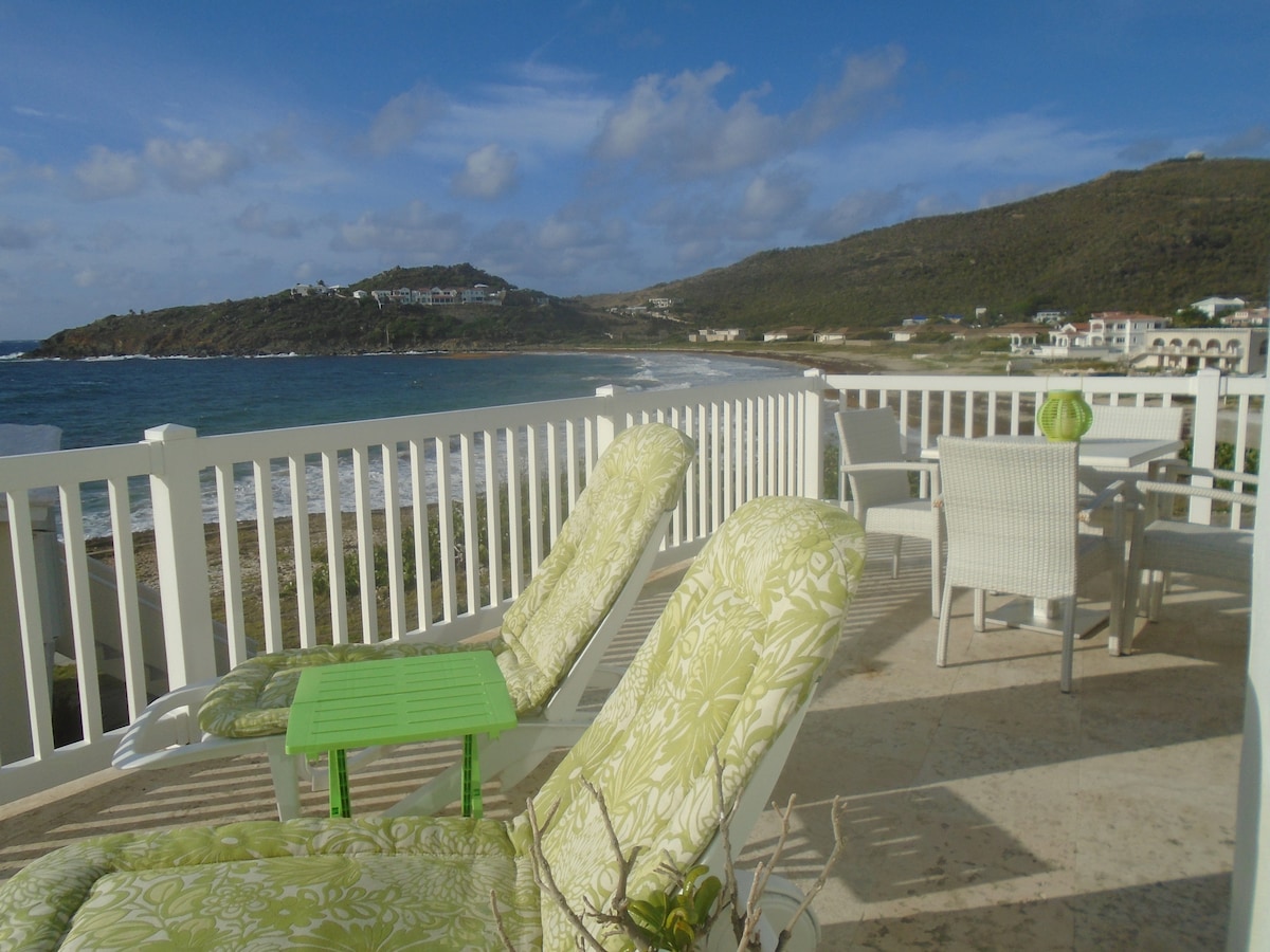 A wrap-around deck features lounge chairs with green floral patterns and a small table. The ocean is visible in the background, with hills rising to either side. The scene is complemented by a partly cloudy sky, enhancing the coastal ambiance.
