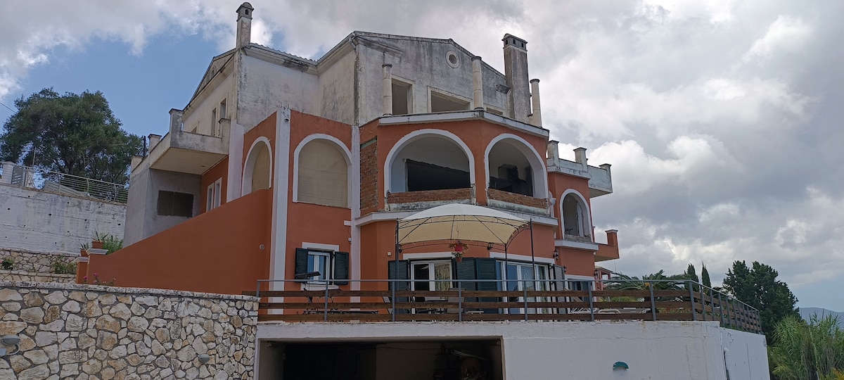 The exterior of a three-story house displays a blend of modern architecture and traditional elements. Arched windows and balconies provide multiple outdoor seating areas. A stone wall surrounds the property, enhancing privacy, while an umbrella shading a patio area adds comfort for outdoor relaxation.