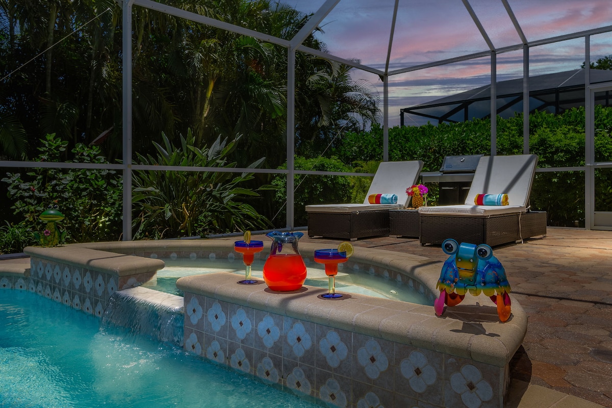A heated saltwater spa is surrounded by a screened lanai, featuring chaise loungers and tropical greenery. Colorful beverages sit on the ledge alongside playful pool toys. The scene is illuminated by the warm glow of the evening sky.