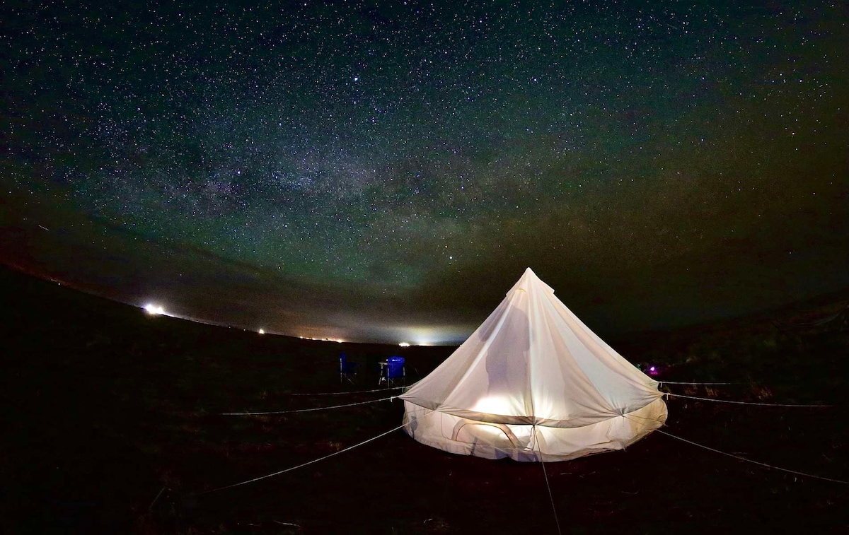 A bell tent stands in an open area under a night sky filled with stars, showcasing the Milky Way. Soft light emits from the entrance of the tent, while two folding chairs are positioned nearby, inviting guests to relax and enjoy the serene atmosphere.