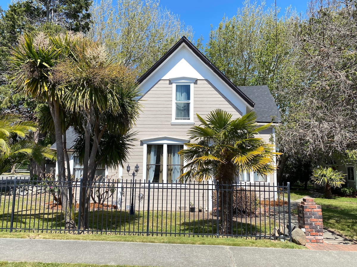 The exterior of a historic Victorian manor is presented, featuring a light-colored facade framed by lush greenery. A fenced yard with palm trees and well-maintained landscaping complements the two-story structure, enhancing its elegant and classic presence. Bright blue skies provide a serene backdrop.
