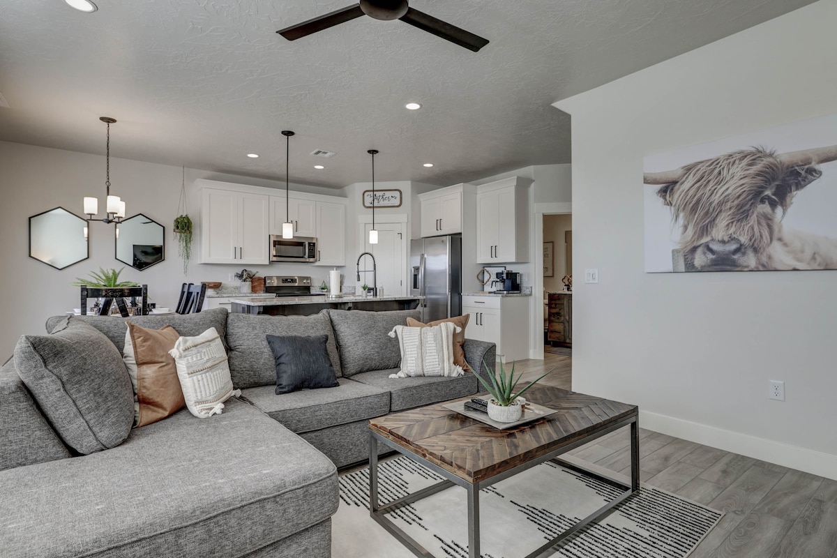The living area features a spacious gray sectional sofa adorned with decorative pillows. A wooden coffee table rests on a black and white patterned rug, complemented by a potted plant. The kitchen area, equipped with modern appliances, is visible in the background.