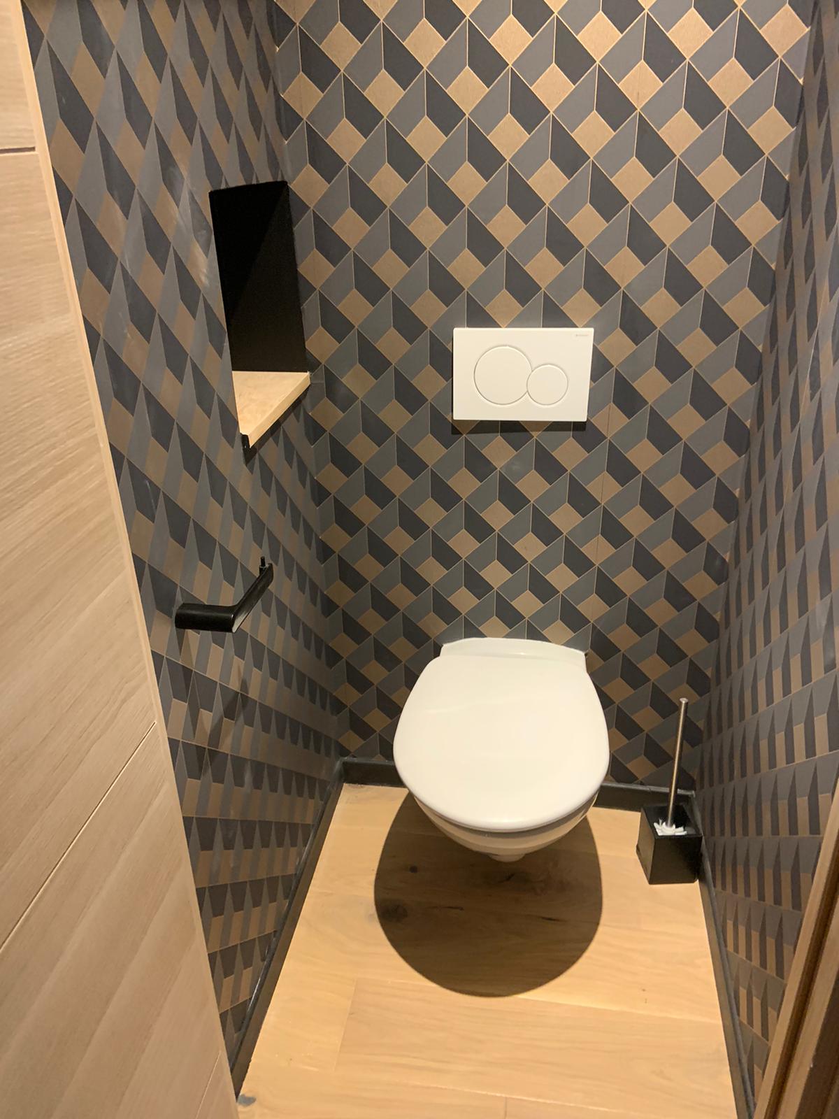 A compact restroom features geometric patterned wallpaper in soft tones. A modern toilet is positioned against a wooden floor. A small shelf is set into the wall for convenience, and a sleek toilet brush holder is visible nearby.