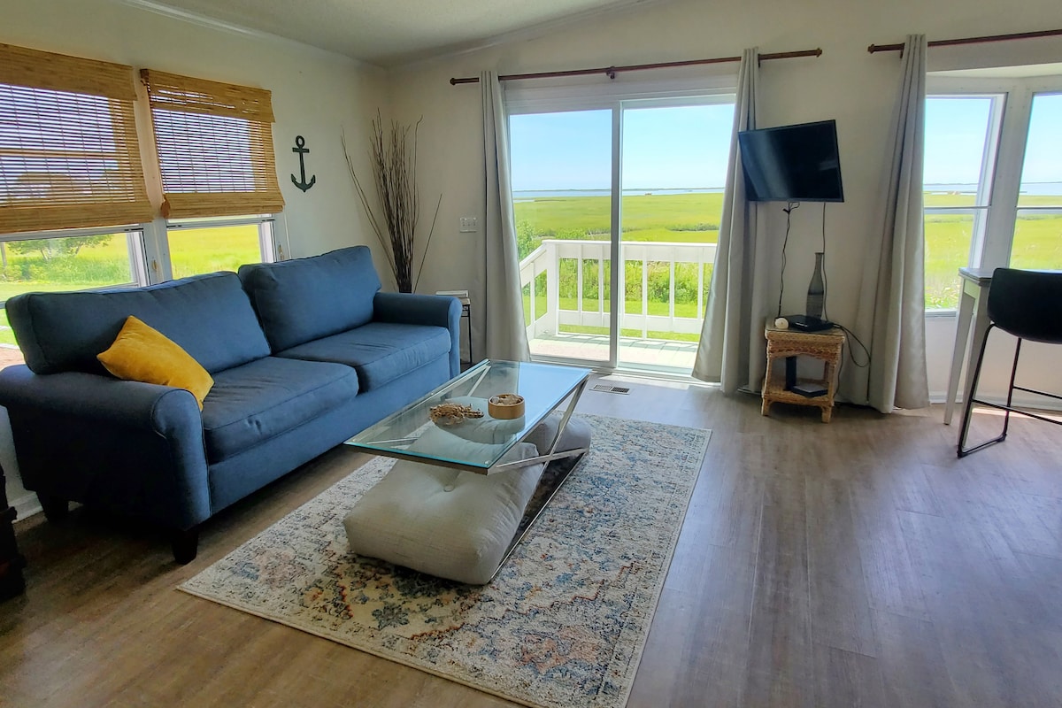 A comfortable seating area features a blue couch with a yellow accent pillow, positioned beside a large window that offers views of the surrounding landscape. A glass coffee table sits atop an area rug, with natural light illuminating the space.