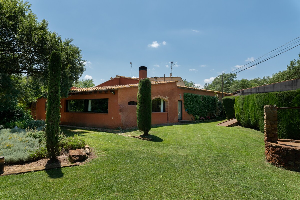 The rustic exterior of the house showcases warm earthy tones under a clear blue sky. Lush green grass surrounds the property, complemented by neatly trimmed hedges and tall conifer trees. Large windows invite natural light, revealing a welcoming atmosphere.