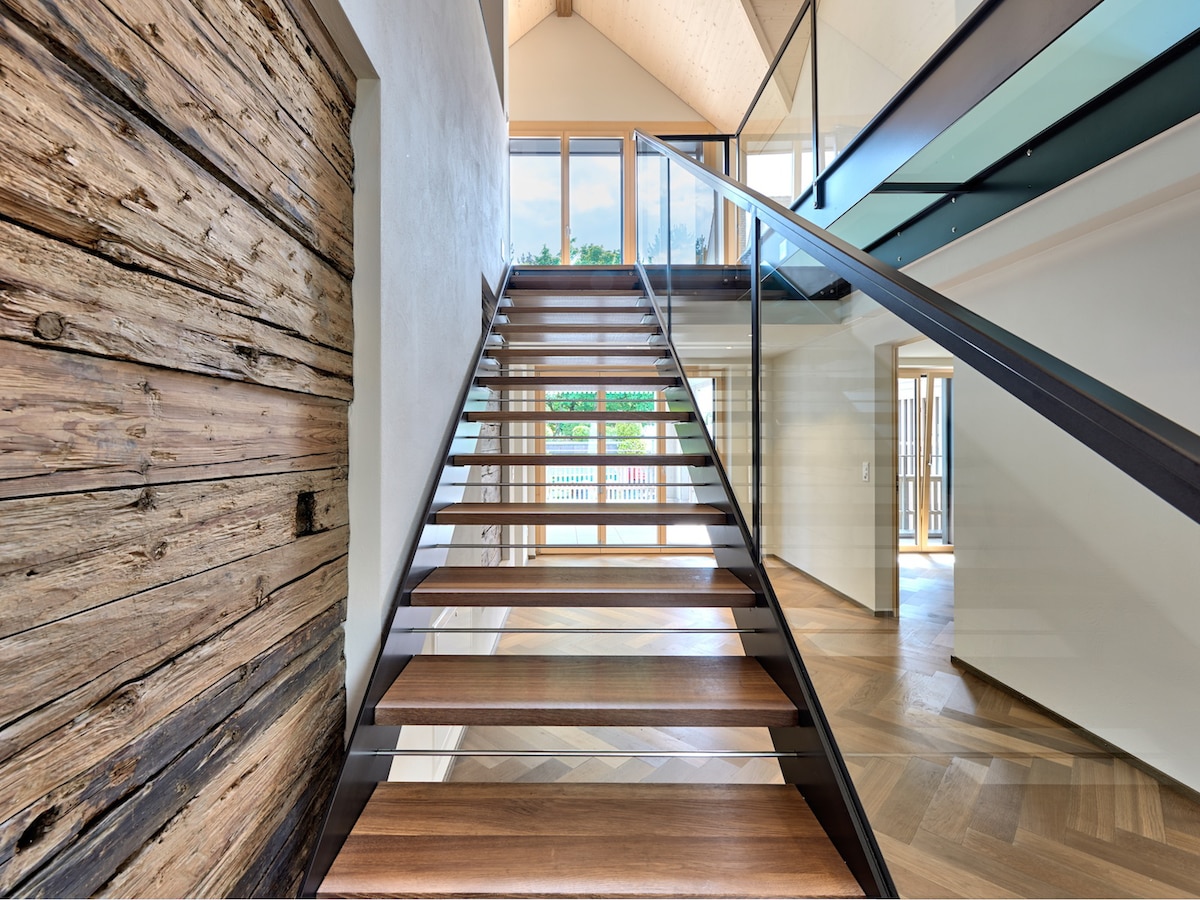 A modern staircase with wooden steps and glass railing ascends to the upper level, complemented by a feature wall of natural wood. Natural light illuminates the space through large windows, providing a view of the surroundings.