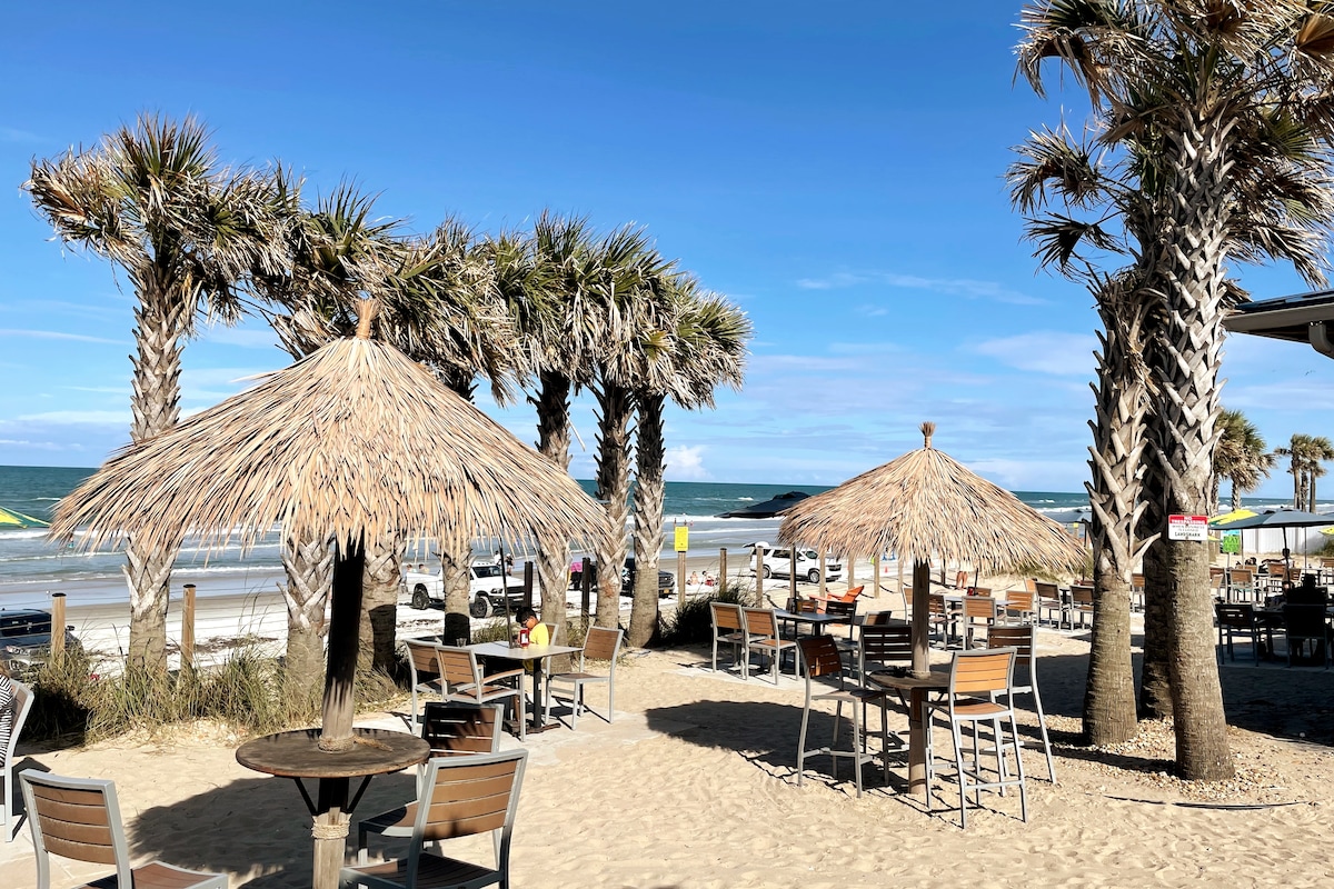 Umbrellas with thatched roofs provide shade over wooden tables and chairs set on a sandy beach. Palm trees frame the scene, with the ocean visible in the background under a blue sky dotted with wispy clouds.