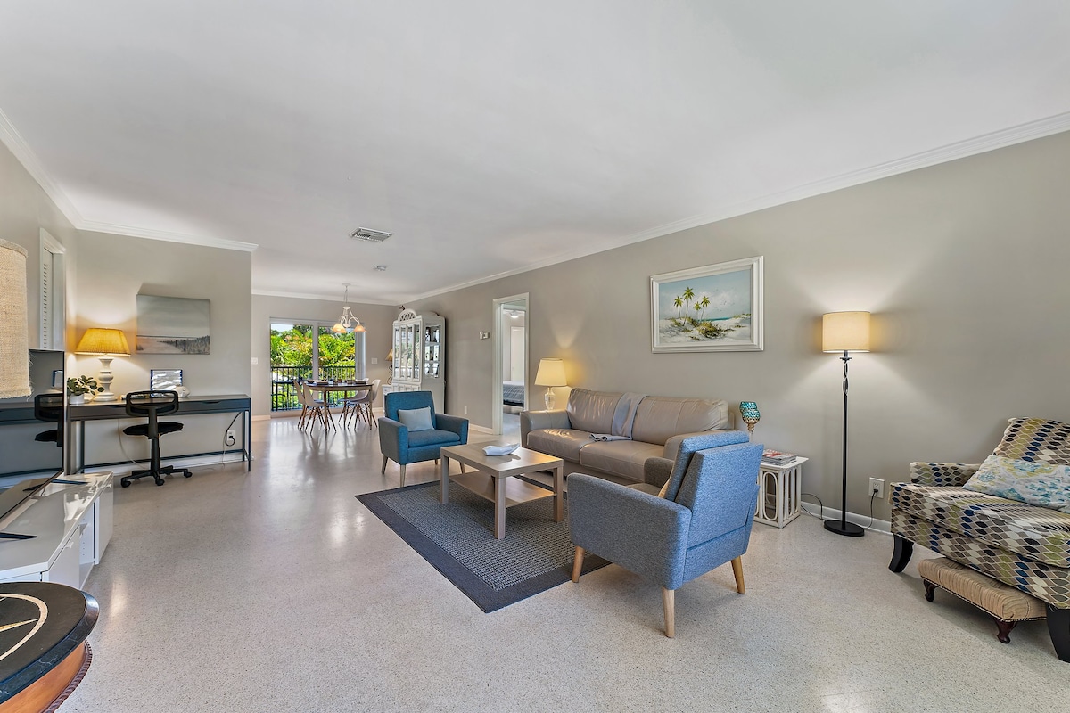 A spacious living area is furnished with a sofa, two armchairs, and a coffee table. A desk with a chair is positioned against the wall, and a large window allows natural light to fill the room. A dining area with a table is visible in the background.