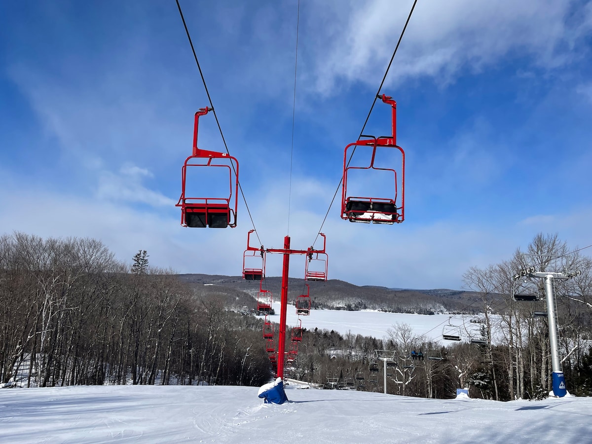 Photo of a ski lift on a clear day, featuring red chairs suspended above a snowy hill. In the background, a serene view of forested slopes and a frozen lake is visible, under a blue sky with scattered clouds.