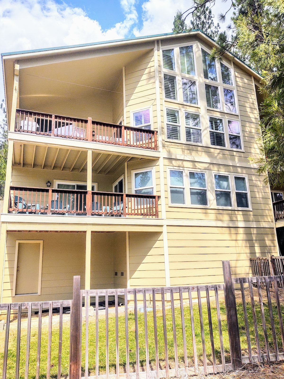 A three-story cabin is showcased, featuring a light yellow exterior with multiple large windows allowing natural light. Two balconies are visible, each with wooden railings. A fenced yard area is situated in front, with green grass enhancing the outdoor space.