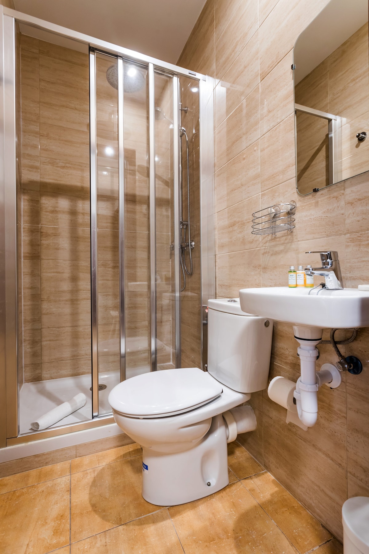 The private bathroom is equipped with a shower featuring glass doors, a toilet, and a sink. Neutral-toned tiles adorn the walls and floor, while a small shelf holds complimentary toiletries. A mirror reflects the natural light within the space.