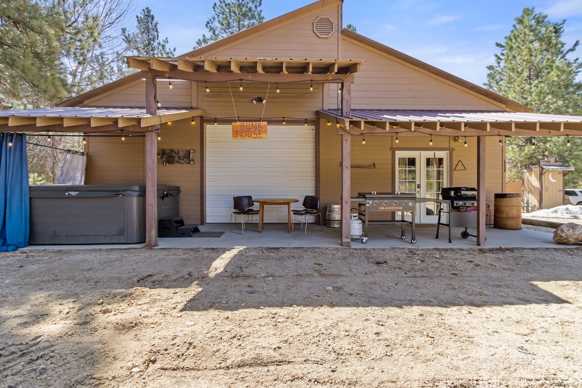 The exterior showcases a spacious covered porch with outdoor seating and a grill. A hot tub is positioned nearby, accompanied by string lights overhead. Surrounding trees provide a natural backdrop, while the sandy ground emphasizes a relaxed outdoor environment.