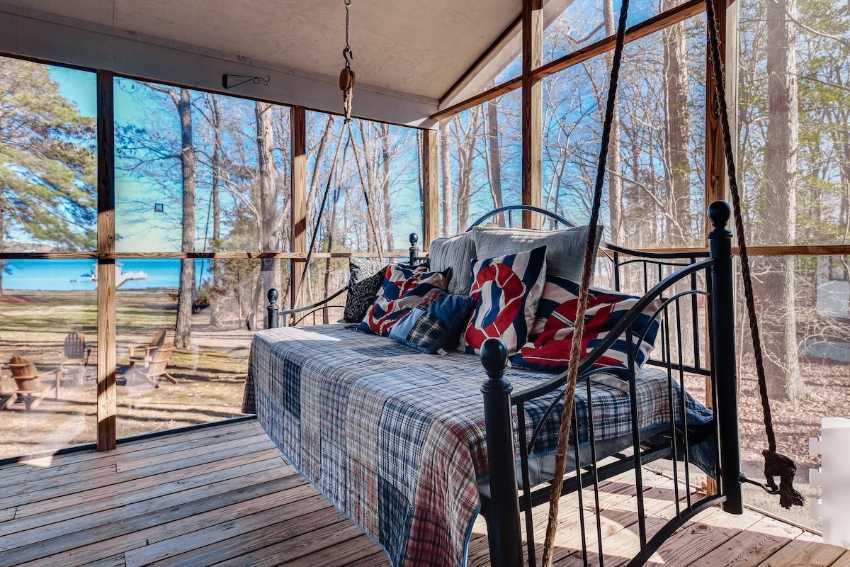 A cozy hanging bed is featured on the screened porch, adorned with various throw pillows. Natural light filters through the screens, revealing a view of the lake and yard beyond, with seating arrangements visible in the outdoor space.