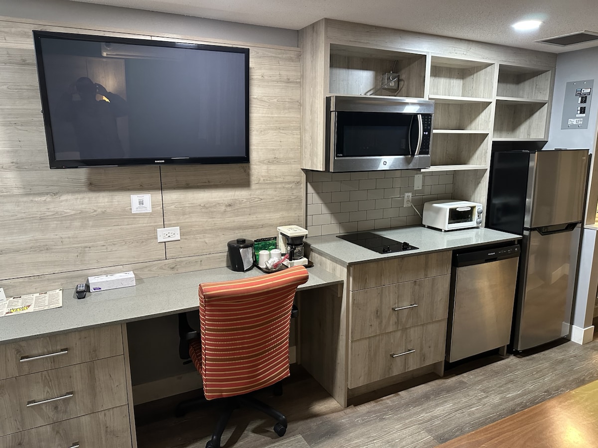 A modern kitchenette is presented with sleek cabinetry and a spacious countertop. Stainless steel appliances include a refrigerator, microwave, and coffee maker. A mounted flat-screen TV is positioned above the counter, while a desk with an orange striped chair offers a workspace.