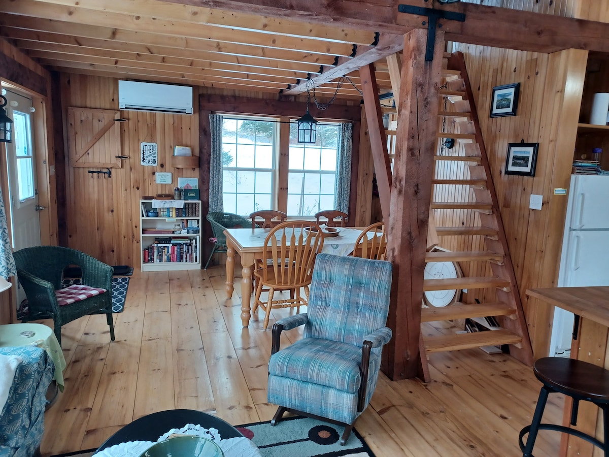 An open interior space is featured, showcasing wooden beams and paneling. A dining table with chairs sits near large windows, while a sitting area includes a plaid armchair and green woven chairs. A staircase leads to an upper level, enhancing the cabin's rustic charm.