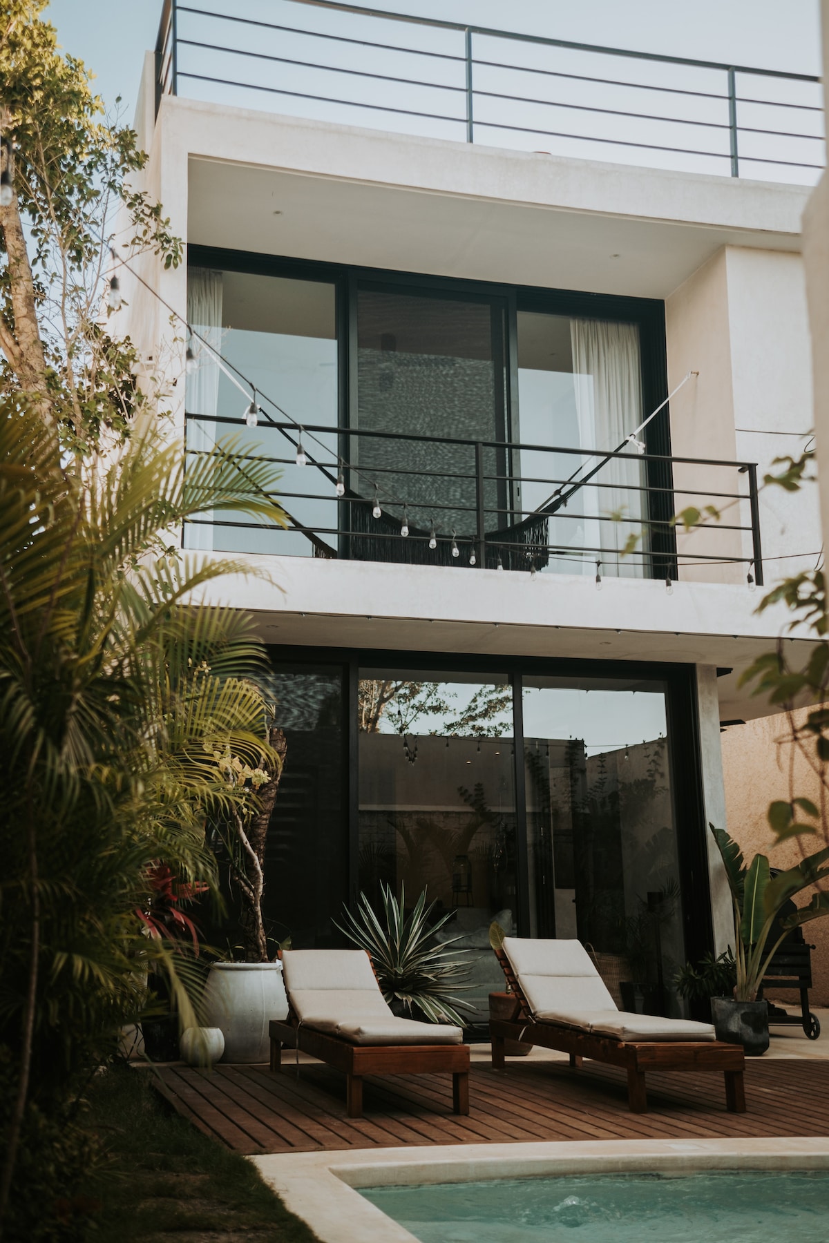 A contemporary villa showcases large glass sliding doors, allowing natural light to fill the space. The outdoor area features two sunbeds positioned beside a tranquil pool, surrounded by greenery and tropical plants, creating a serene retreat atmosphere.