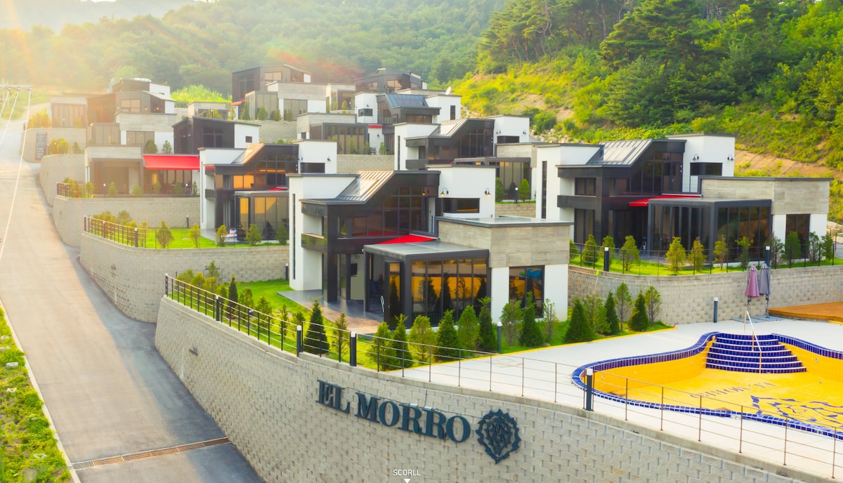 A collection of modern villas is arranged on a hillside, featuring sleek architecture with large windows and landscaped gardens. A distinctive pool area is visible, along with colorful awnings that provide shade. The surrounding hills create a serene backdrop for the property.