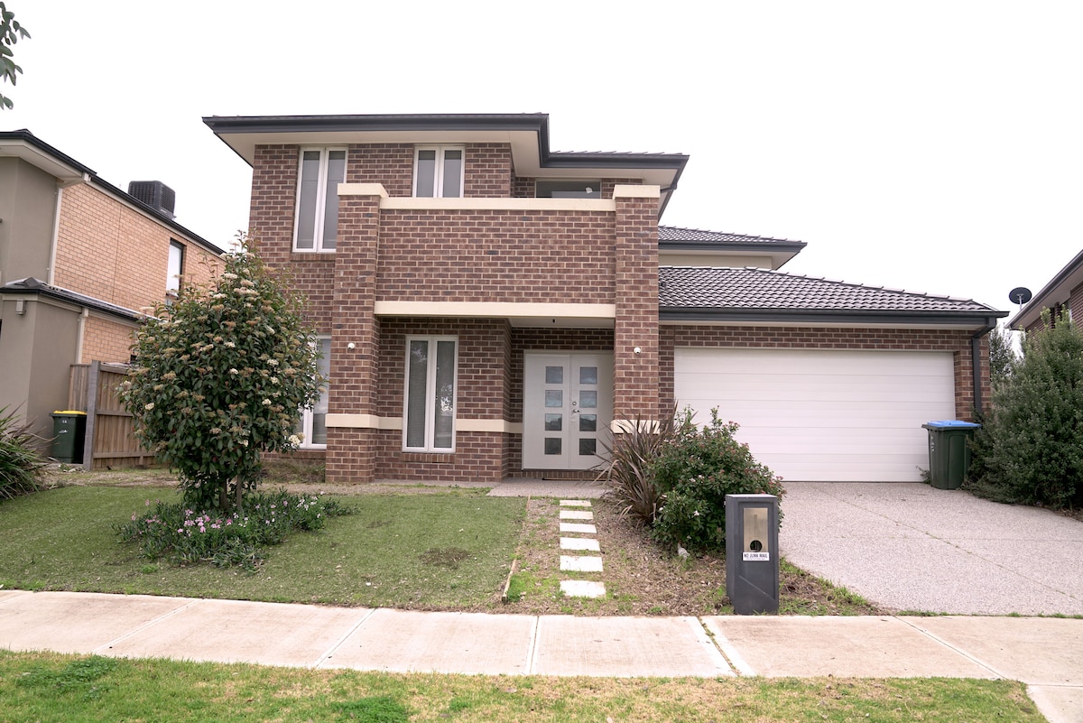 The exterior of a modern two-story house features a brick façade with large front windows. A neatly landscaped yard surrounds the entrance, which is flanked by shrubs. A two-car driveway leads to a garage door, with a blue bin positioned nearby.