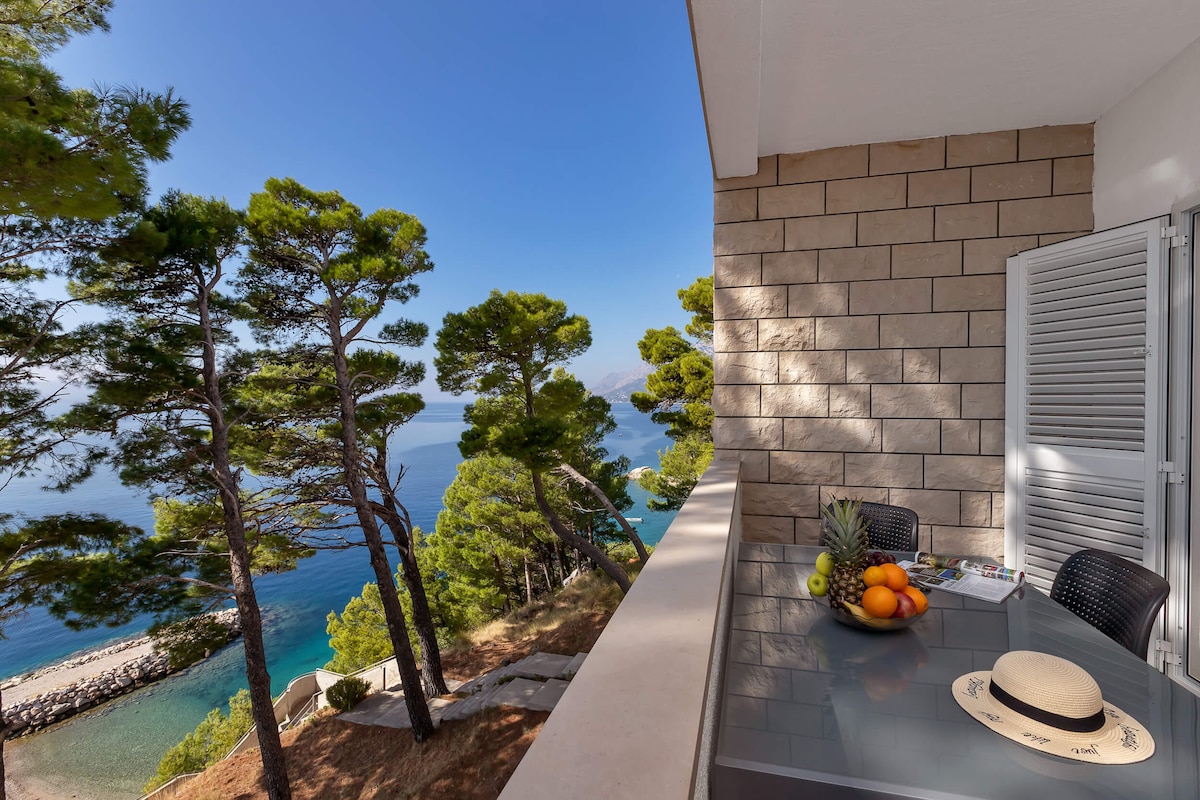 An outdoor balcony is framed by tall pine trees, offering a view of the sea and nearby pebble beach. A modern table is set with a bowl of colorful fruit, while a wide-brimmed hat rests nearby. Bright sunlight illuminates the serene, natural surroundings.