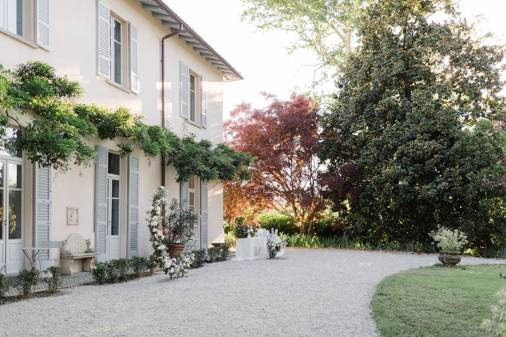 A charming exterior of a villa is presented, surrounded by lush greenery and flowering plants. The gravel pathway leads to a beautifully manicured lawn. Sunlight filters through trees, casting dappled shadows on the building's soft-colored walls, highlighting the elegant windows adorned with shutters.
