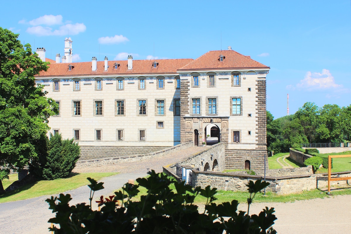 An elegant historic building stands against a blue sky, showcasing its light-colored stone façade and distinctive red roof. Lush greenery surrounds the structure, with a pathway leading to an arched entrance. The scene conveys a sense of tranquility and heritage.