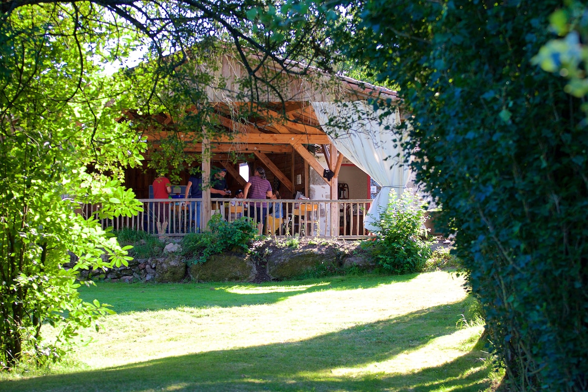 A cabin is set amidst lush greenery, showcasing a spacious covered terrace framed by wooden beams. The open area features outdoor seating for enjoying nature, with grassy surroundings and trees creating a serene atmosphere. Natural light filters through the foliage, enhancing the inviting environment.