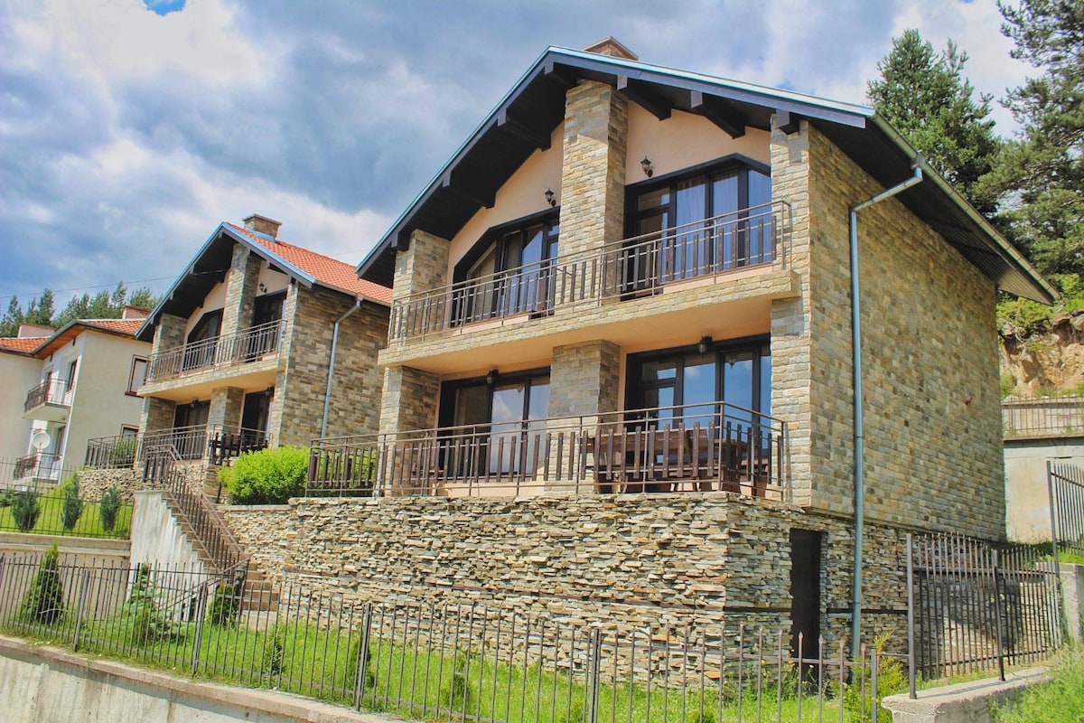 A spacious stone house is prominently displayed, featuring multiple levels and large glass doors that offer views of the surrounding landscape. A balcony with railings extends from the upper level, enhancing the exterior's charm and inviting fresh mountain air.