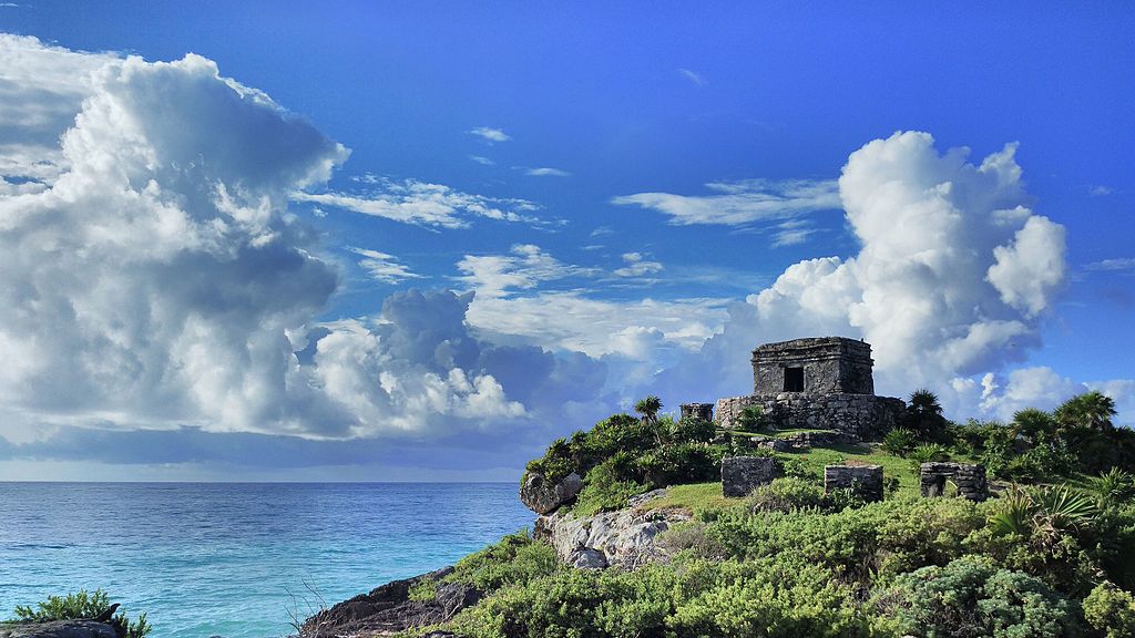 An ancient stone structure is positioned on a rocky outcrop overlooking the ocean. Lush greenery surrounds the site, with the blue waters of the sea visible below. A bright sky filled with fluffy clouds creates a serene backdrop for this historic location.