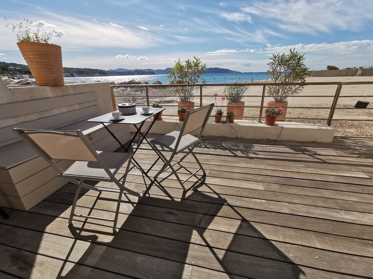 A spacious terrace features a small table and two folding chairs, complemented by potted plants. The serene view includes the sea and the sky, creating a tranquil outdoor space for relaxation.