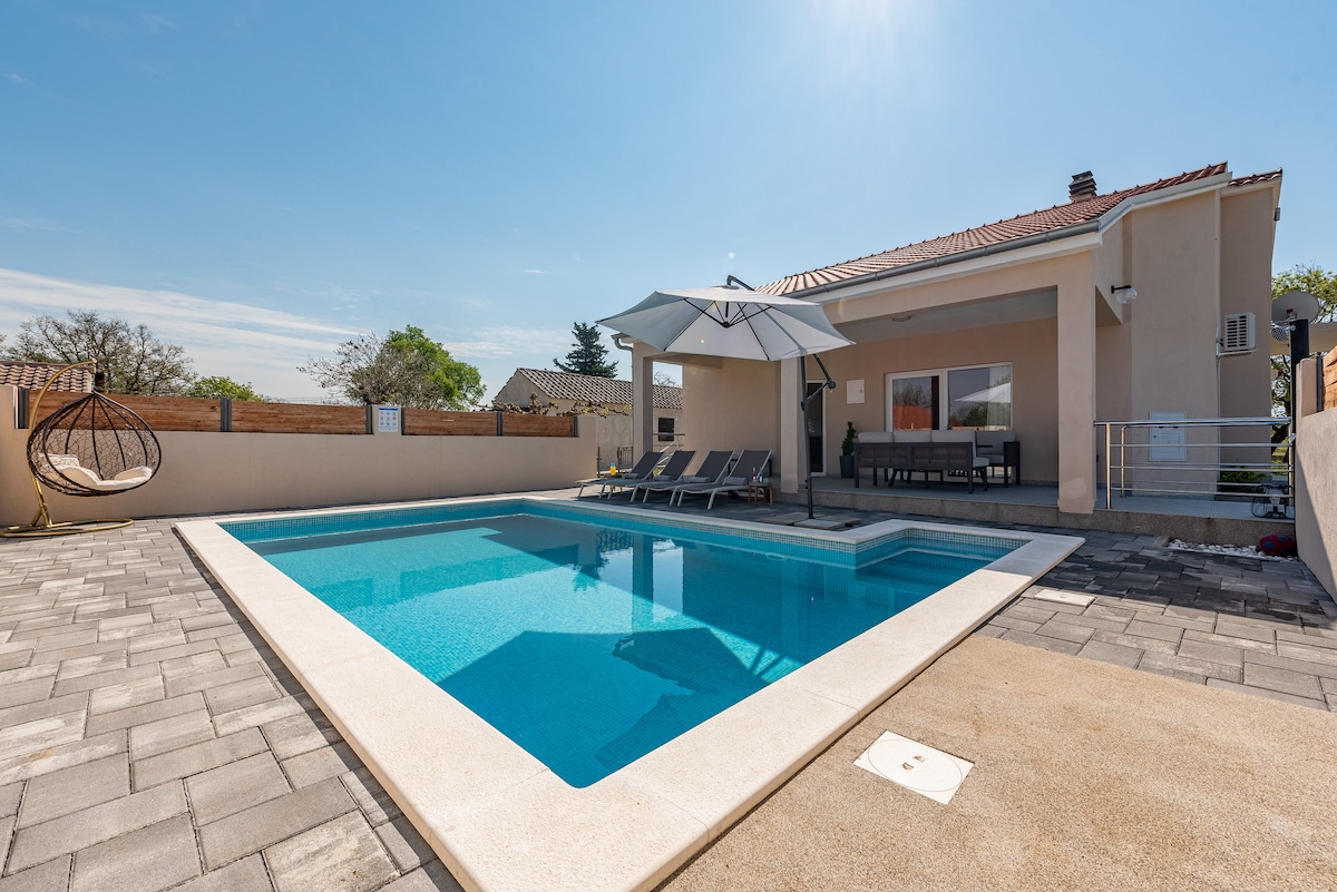 The image showcases a clear, inviting swimming pool surrounded by a stone patio. Sun loungers are arranged around the pool, and a large umbrella provides shade. A dining area with outdoor seating is visible near the house, complemented by a well-maintained fence and greenery in the background.