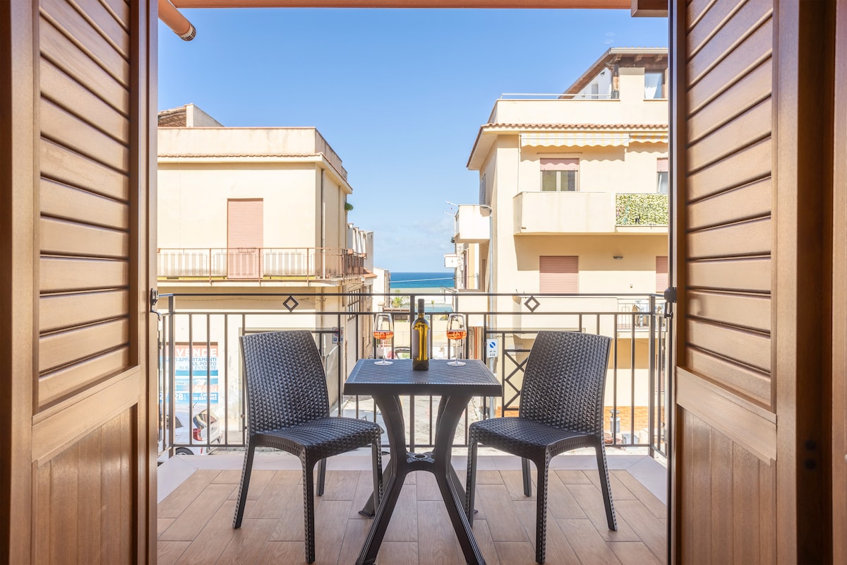 A private balcony is framed by open shutters, featuring a table and two chairs. The view includes nearby buildings and a glimpse of the sea, creating an inviting outdoor space for relaxation.