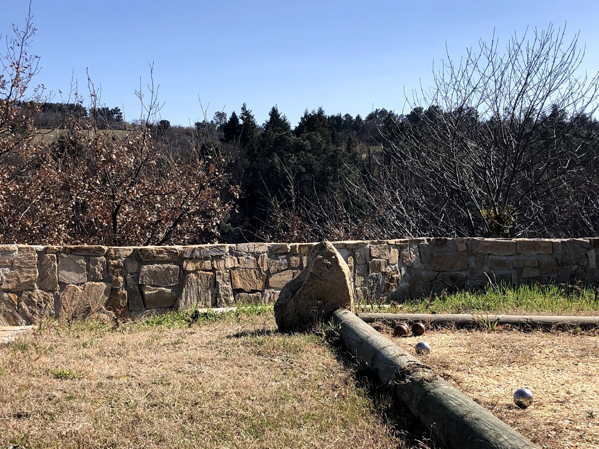 A natural outdoor area features a stone wall and a grassy field, with a large rock placed prominently at the forefront. Metal balls, used for pétanque, are scattered on the ground. In the background, trees provide a glimpse of the surrounding landscape.
