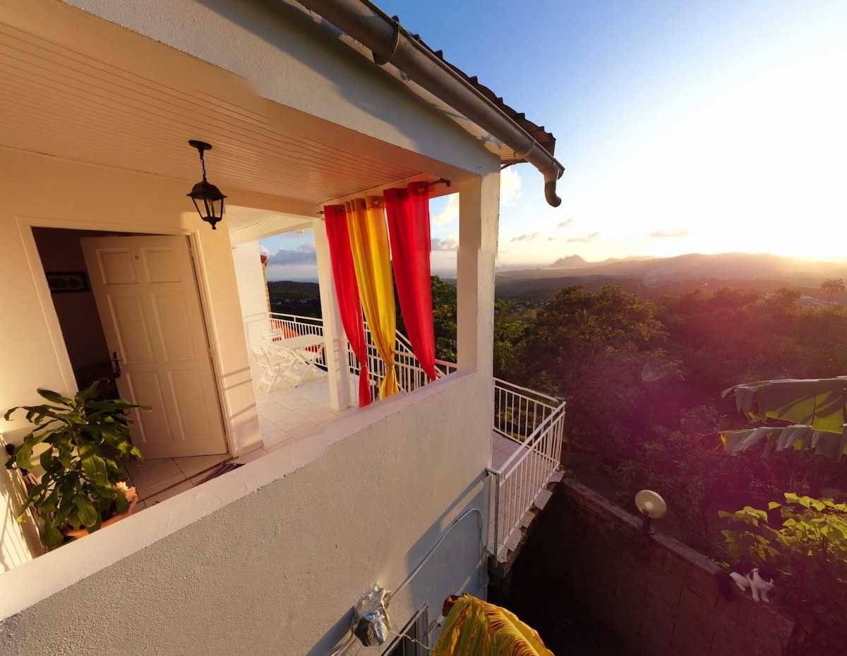 A spacious terrace features colorful curtains flowing in the breeze, framing a panoramic view of the lush landscape below. Potted plants add greenery, while a table and chairs are positioned for relaxation. The setting sun casts a warm glow over the scene.