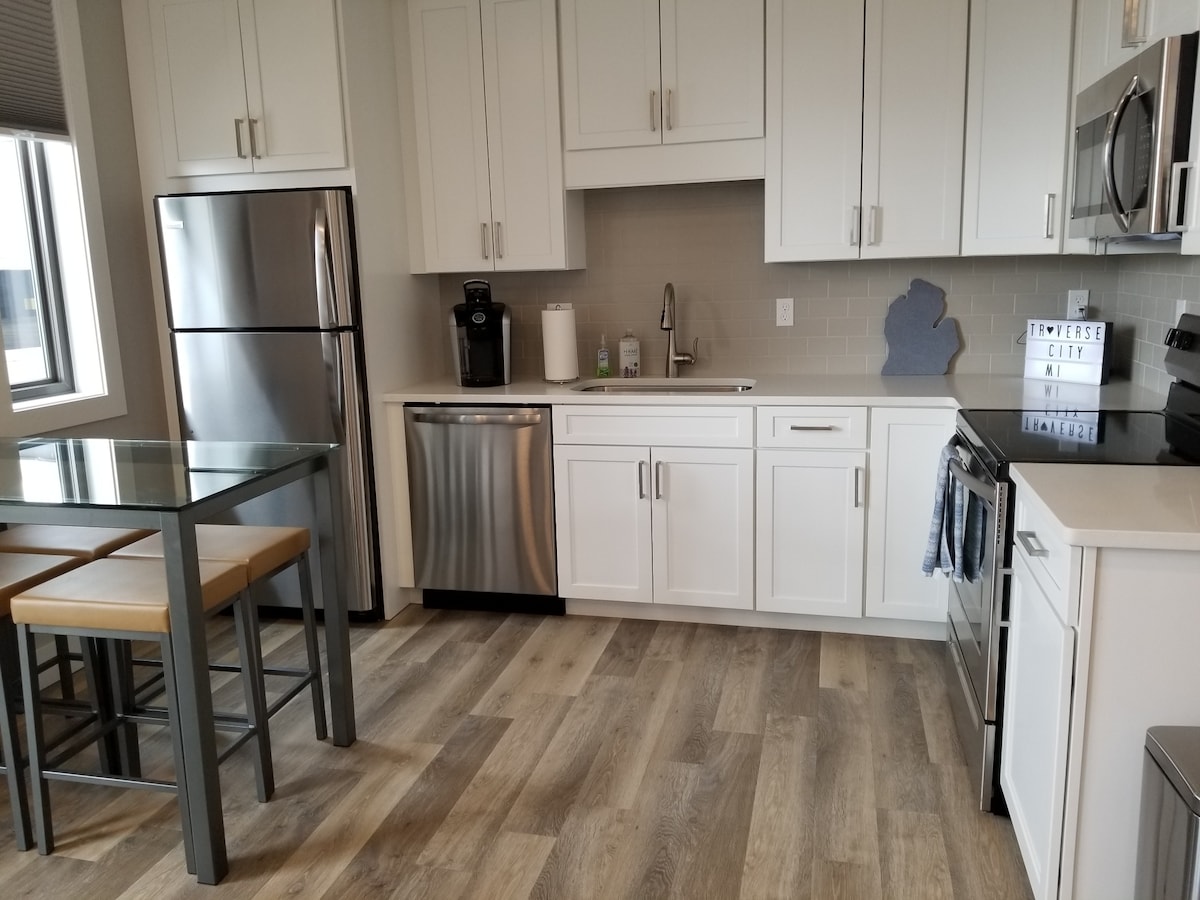 The kitchen features modern stainless steel appliances, including a refrigerator, dishwasher, and oven. A small dining table with four chairs is positioned near a window, creating a bright and functional space. Light wood flooring enhances the contemporary design.