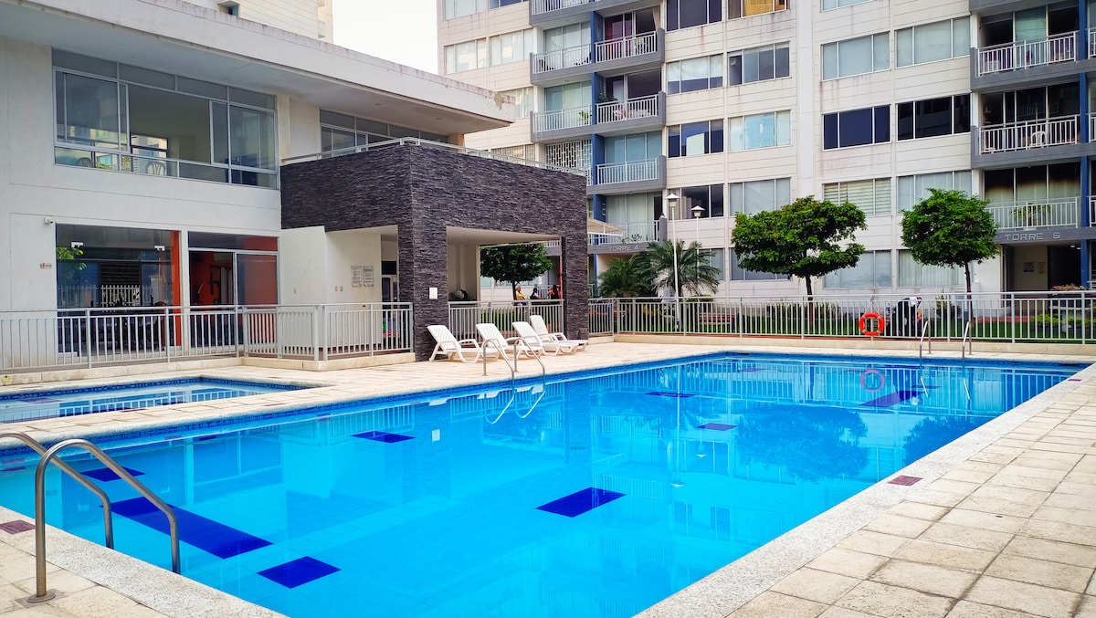 An outdoor pool is surrounded by a clean and organized patio area. Comfortable lounge chairs are positioned beside the pool, and the building’s modern architecture creates a welcoming environment. Lush greenery and well-maintained landscaping are visible in the background.
