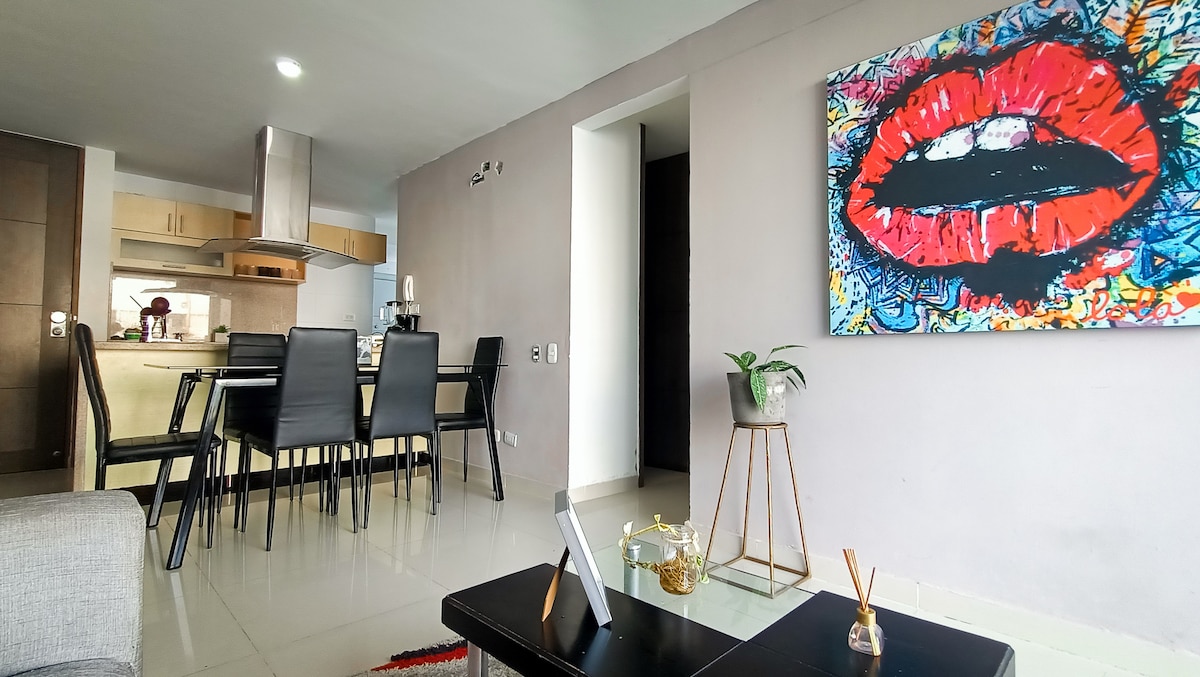 A modern living space is showcased with a dining area featuring a rectangular glass table surrounded by four black chairs. A colorful wall art piece depicting red lips is displayed prominently. Natural light fills the room, reflecting off the glossy floor tiles.