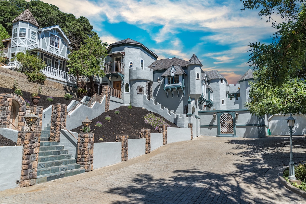An enchanting castle-like structure stands amidst lush greenery, characterized by its light gray exterior and turret-style roof. The exterior includes multiple levels with intricate architectural details, complemented by a paved driveway and manicured landscaping that enhances the overall charm of the property.