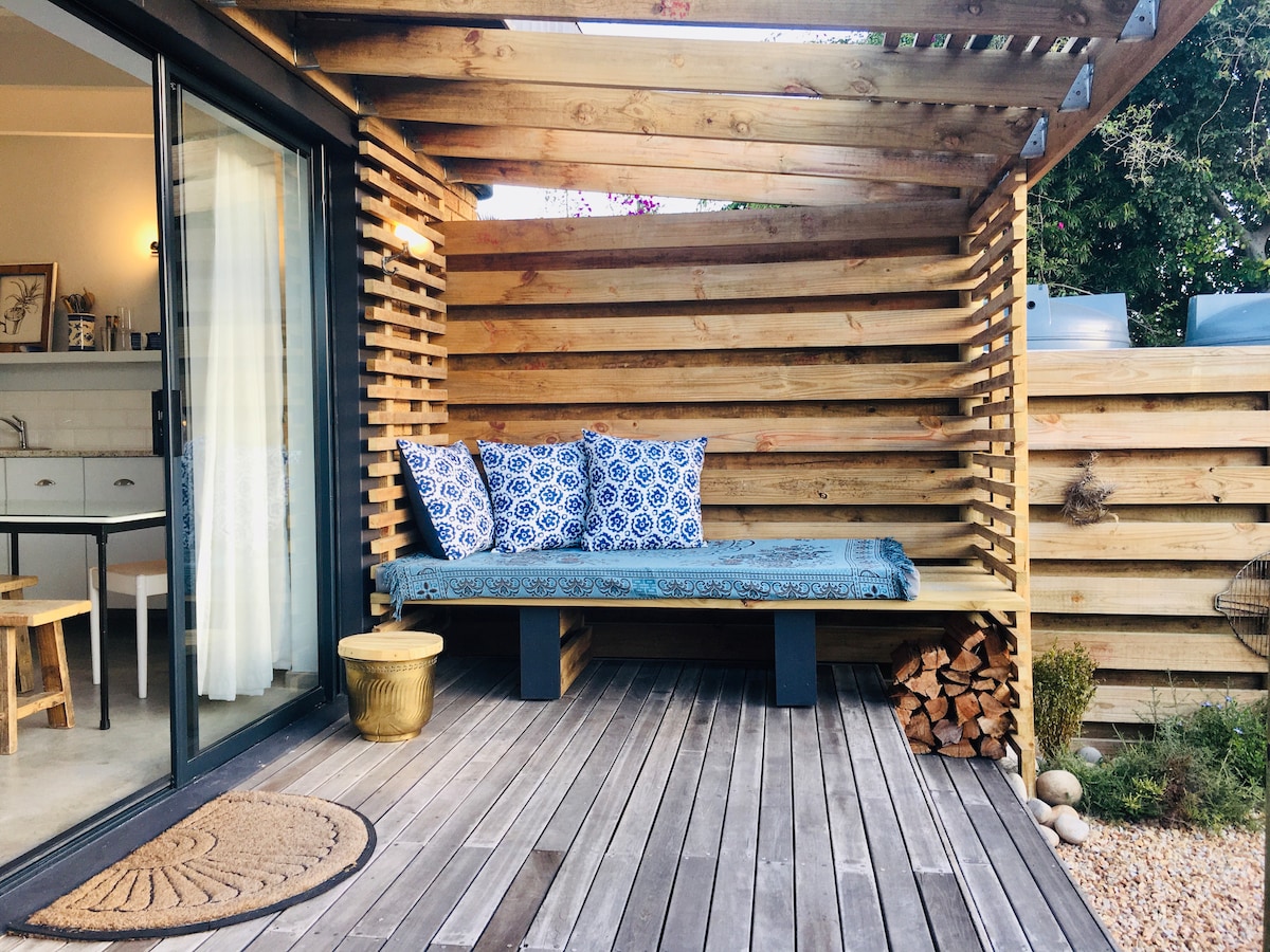 A wooden deck area features a comfortable seating nook adorned with blue patterned cushions. Soft lighting enhances the inviting atmosphere. A wooden slatted fence provides privacy, while a small gold table complements the space. The background showcases a modern kitchen with light-colored cabinetry.