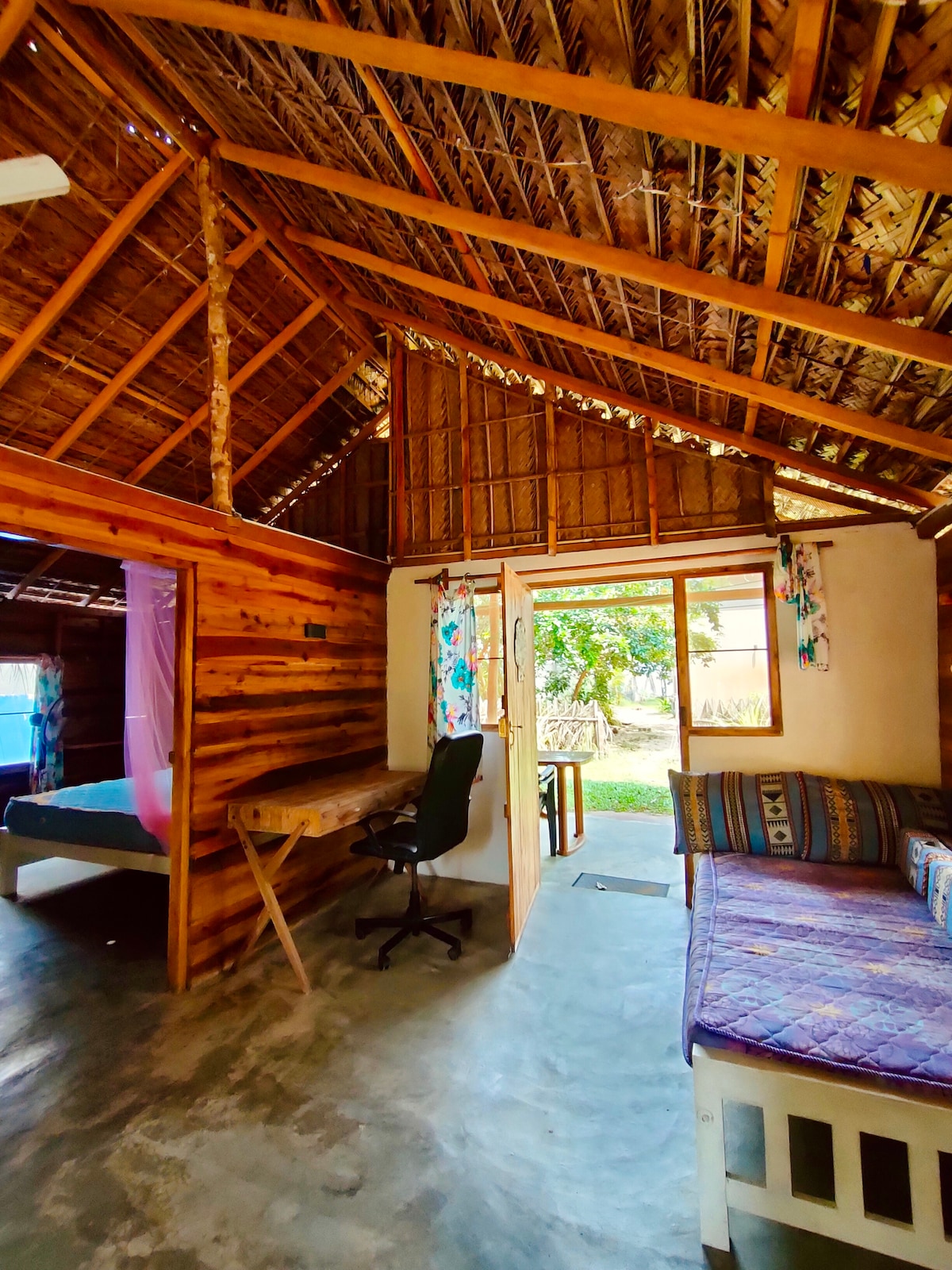 An interior view of a rustic cottage showcases wooden walls and a high thatched ceiling. A spacious bed and a cozy sitting area with a colorful cushion can be seen. A desk with a chair is positioned near an open doorway that leads to a garden area.