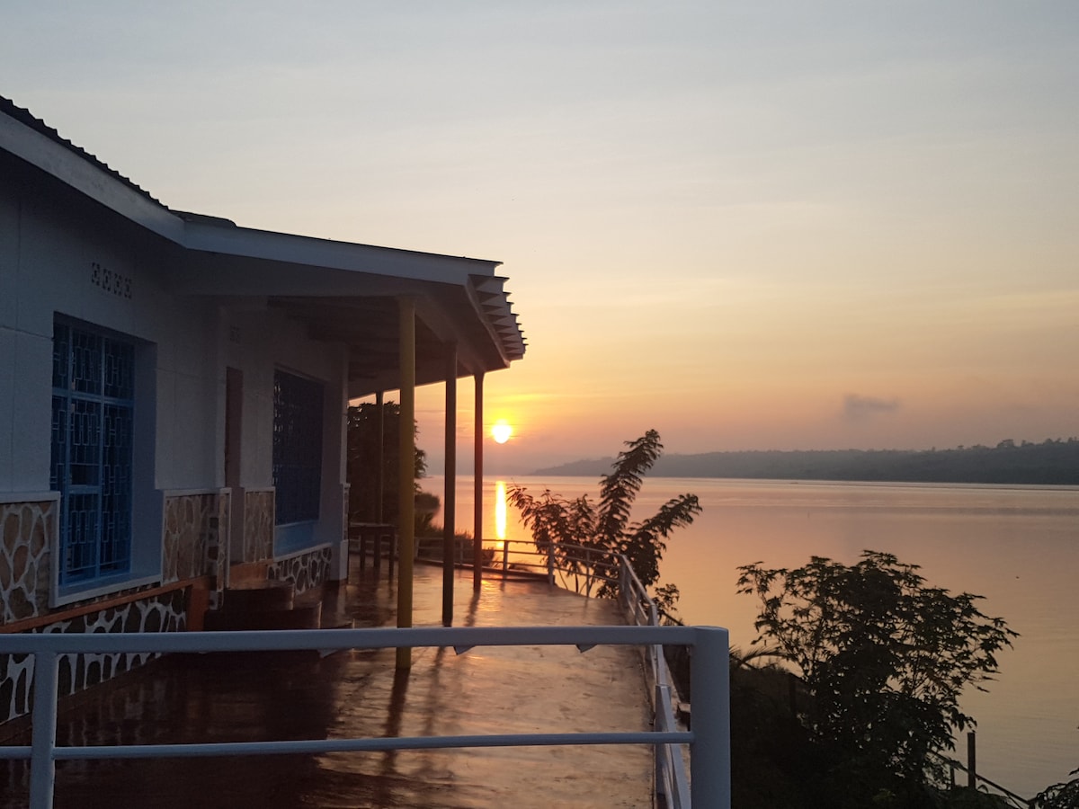 A serene view from the porch captures the sun rising over the lake, reflecting softly on the water's surface. The building features blue windows and wooden accents, framed by lush greenery and a peaceful shoreline.