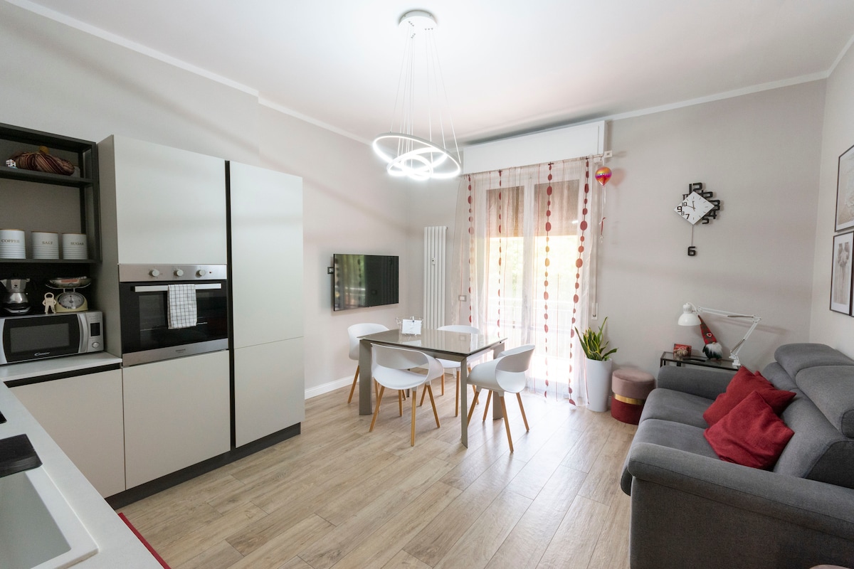 A modern kitchen is equipped with sleek cabinetry and appliances, including a microwave and oven. A dining table with white chairs is positioned next to a comfortable sofa adorned with cushions. Natural light filters through sheer curtains, enhancing the welcoming ambiance.