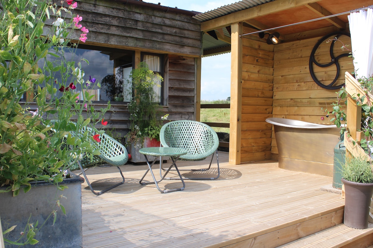 A spacious outdoor deck features comfortable seating and a luxurious copper bath. Surrounding greenery adds a natural touch, while a rustic wood paneling complements the cabin's exterior. The setting invites relaxation with views over open fields.