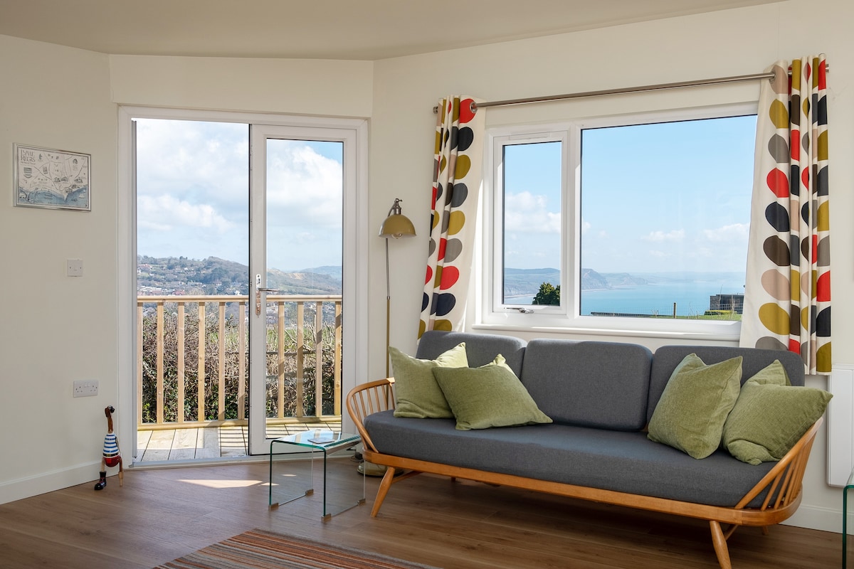 A modern lounge area features a comfortable gray sofa adorned with green cushions. Large windows provide expansive views of the sea and surrounding landscape, with natural light illuminating the space. A small glass table complements the decor, framed by colorful patterned curtains.