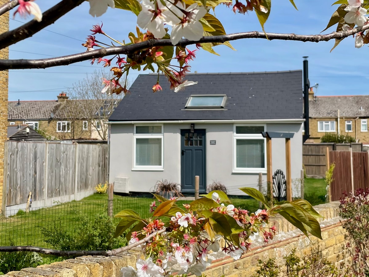 The cottage's exterior is framed by blooming flowers, showcasing a modern, single-story design with a welcoming dark front door and a small window above. Green grass surrounds the property, complemented by a fenced garden area. A wood-burning stove chimney is visible on the roof.