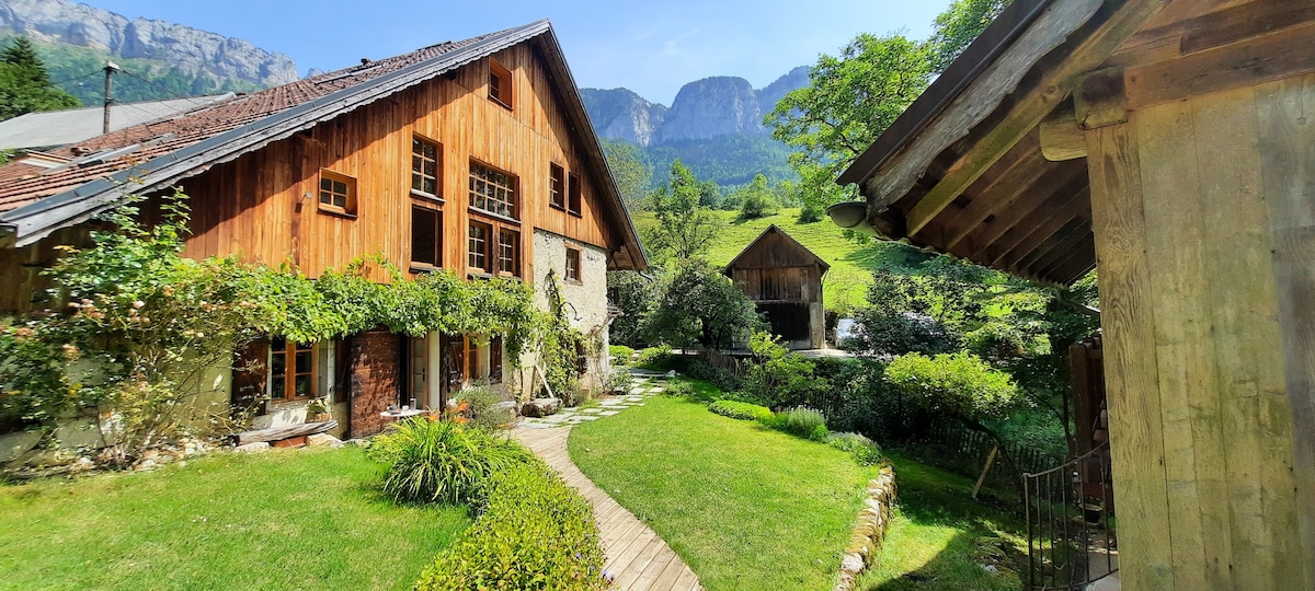 A rustic chalet is surrounded by lush greenery and vibrant landscaping. Large windows reveal wooden interiors, while a pathway leads through the garden toward additional structures. The backdrop features majestic mountains, contributing to the tranquil alpine setting.