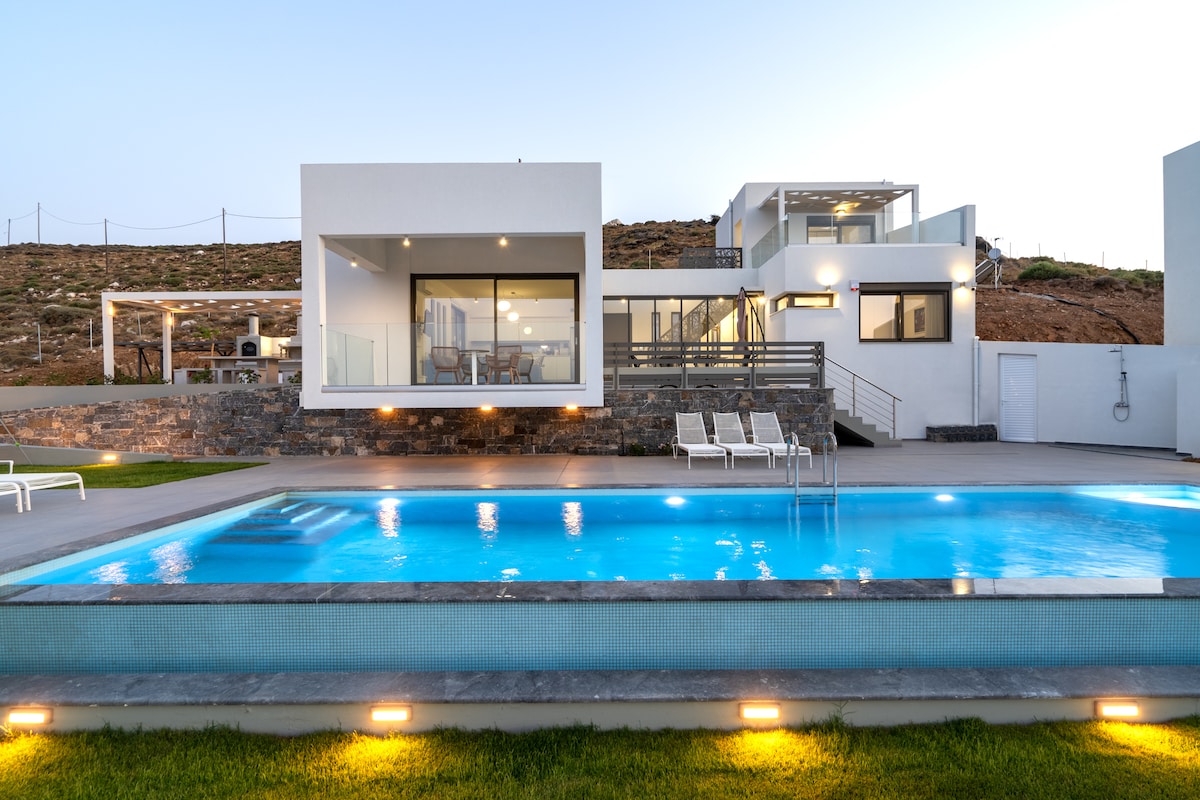 A modern villa is showcased with a spacious terrace and an inviting outdoor pool. Sun loungers are positioned beside a clear blue pool, reflecting the evening sky. The structure features large glass windows that offer views of the interior dining area and surrounding landscape.
