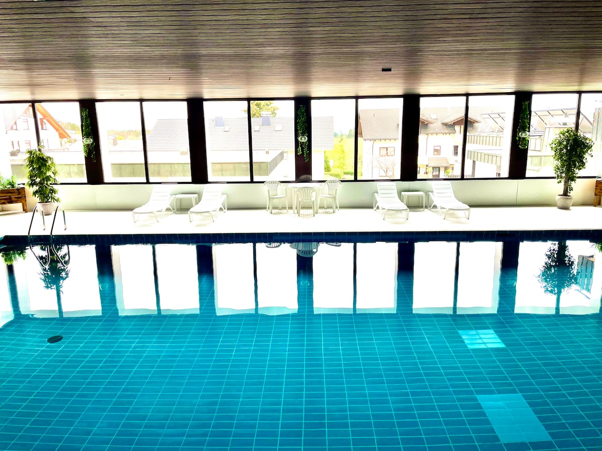 A spacious indoor swimming pool is surrounded by large windows with a view of the outdoors. Comfortable lounge chairs are placed around the pool area, and potted plants provide a touch of greenery. The water's reflection creates a calming atmosphere.